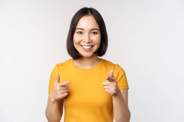 Genç Koreli kız öğrenci, kamerayı işaret ediyor ve gülümsüyor, seni tebrik ediyor, insanları seçiyor, beyaz arka planda duruyor..