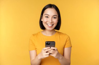 Teknoloji. Gülümseyen Asyalı kadın cep telefonu kullanıyor, akıllı telefonu elinde tutuyor, sarı arka planda tişörtle dikiliyor..