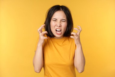Kızgın Asyalı kadın görüntüsü, bağırıyor ve küfür ediyor, öfkeli bir yüz ifadesi, sarı arka planda duruyor..