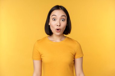 Şaşırmış görünen Asyalı bir kız görüntüsü, şaşırmış, şaşırmış, kaşlarını kaldırmış, sarı arka planda duruyor..