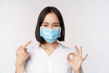 Tıbbi maske takan Asyalı kızın portresi iyi olduğunu gösteriyor ve beyaz arka planda duran kapalı kapılı korumasına işaret ediyor. Boşluğu kopyala