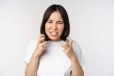 Kızgın Asyalı kadın küfür ediyor, öfkeli ve sinirli görünüyor, dişlerini sıkmış ve kaşlarını çatmış, beyaz arka planda duruyor..