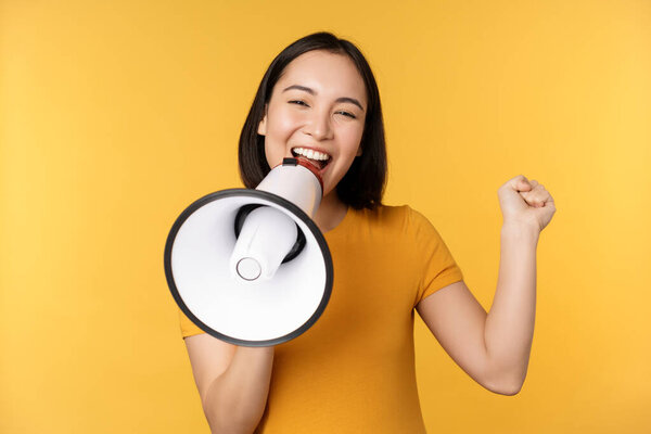 Smiling asian woman standing with megaphone, announcing smth, advertising product, standing over yellow background.