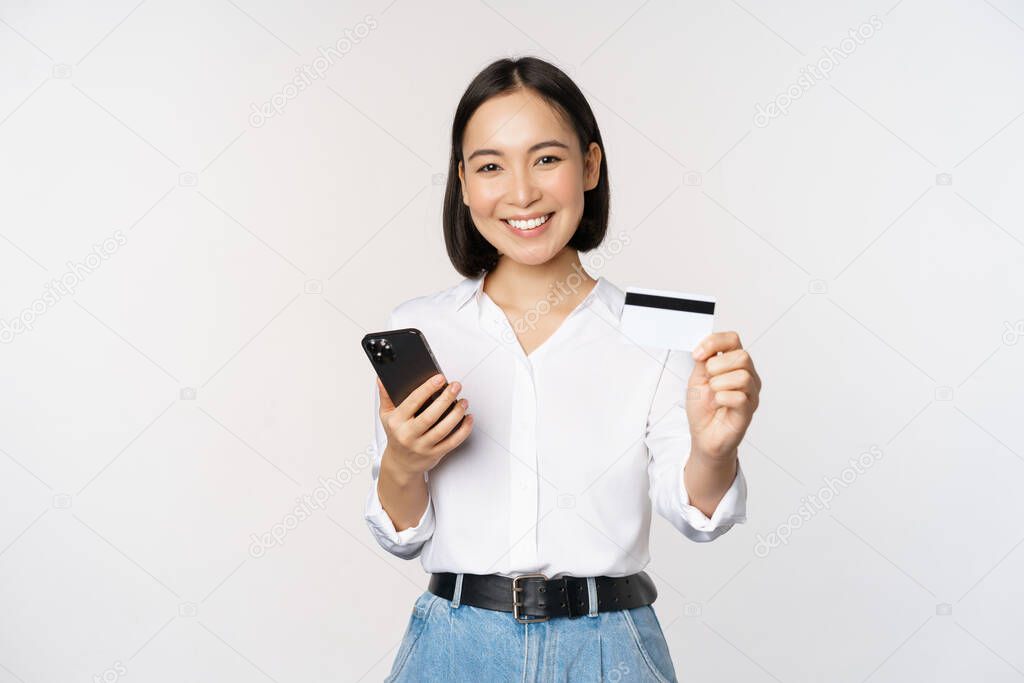 Online shopping concept. Smiling modern asian girl shows her credit card, holds mobile phone, order with smartphone, standing over white background.