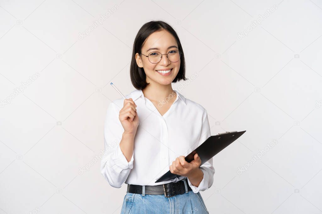 Smiling young asian woman taking notes with pen on clipboard, looking happy, standing against white background.