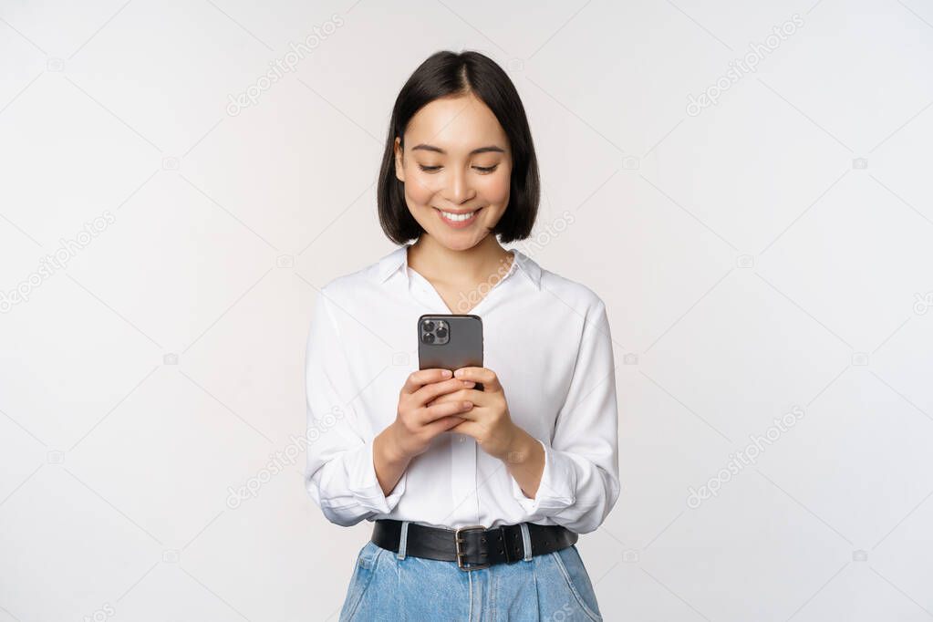 Stylish modern asian girl using mobile phone application, chatting on cellphone and smiling, standing in white blouse against studio background.