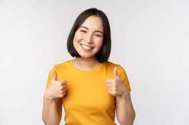 Güzel gülümseyen Asyalı kadın model, başparmaklarını kaldırıyor ve memnun görünüyor, tavsiye ediyor, olumlu geri bildirim yapıyor, beyaz arka planda duruyor..