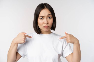 Genç Asyalı kadın kendini inançsızlıkla işaret ediyor, seçiliyor, adaylığı karşısında şaşırıyor, beyaz arka planda duruyor..