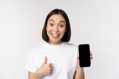 Hevesli genç bir kadın başparmağını kaldırıyor ve cep telefonu ekranında beyaz arka planda tişörtle dikiliyor. Boşluğu kopyala
