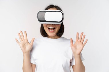Mutlu Asyalı kadın VR kulaklık kullanıyor, el sallıyor ve gülüyor, sanal gerçeklik gözlüğü kullanıyor, beyaz arka planda duruyor..
