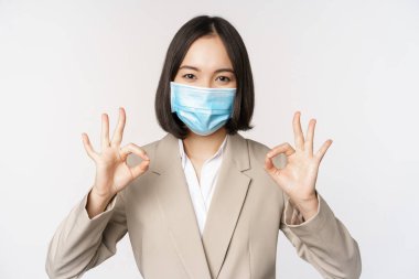 Coronavirus ve işyeri konsepti. Asyalı bir satıcı, tıbbi maske takan bir şirket çalışanı. İşaret veriyor, gülümsüyor, mutlu, beyaz bir geçmişi var..