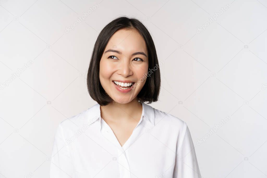 Image of smiling asian woman planning, thinking of smth, daydreaming, standing over white background with smug face. Copy space