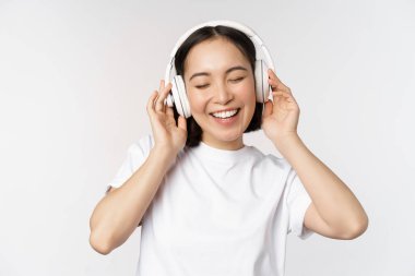 Modern Asyalı kız dans ediyor, kulaklıkla müzik dinliyor, mutlu gülümsüyor, beyaz arka planda tişört giyiyor..