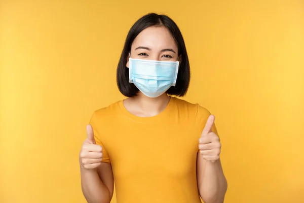 医者の顔のマスクでアジアの女性の笑顔 親指の承認を表示 のように スミスをお勧めします 黄色の背景の上に立って — ストック写真