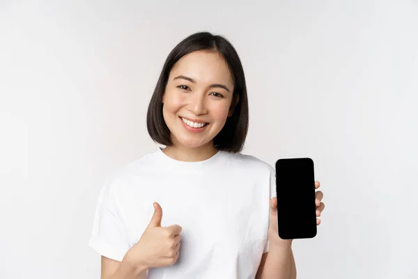 Портрет красивої азіатки, що показує великий палець і екран смартфона, посміхається під час реклами мобільного додатку, білого фону — стокове фото