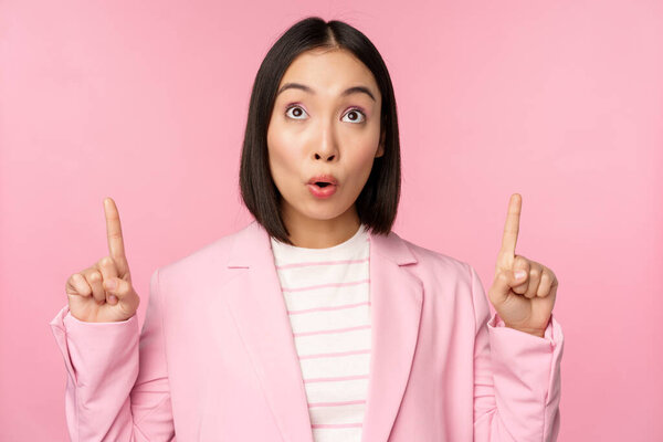 Enthusiastic corporate worker, asian business woman pointing fingers up and smiling, showing advertisement, logo, standing over pink background
