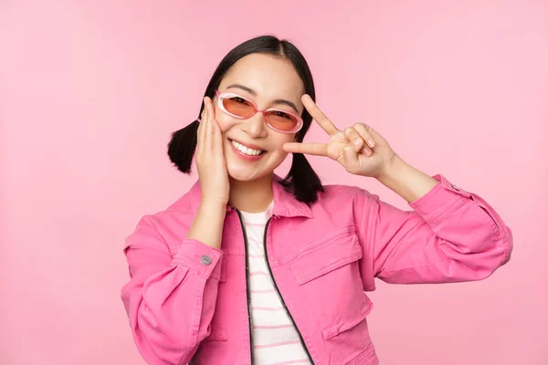 Πορτρέτο του Kawaii ασιατικό κορίτσι με γυαλιά ηλίου, που δείχνει την ειρήνη, v-σημάδι κοντά στο μάτι και χαμόγελα χαριτωμένο στην κάμερα, ποζάρουν σε μοντέρνα ρούχα σε ροζ φόντο — Φωτογραφία Αρχείου