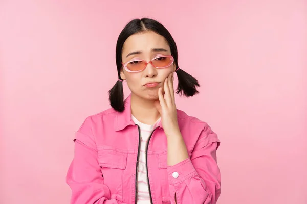 Immagine di triste ragazza asiatica imbronciato, toccando la guancia, il broncio deluso, ha mal di denti, in piedi su sfondo rosa — Foto Stock
