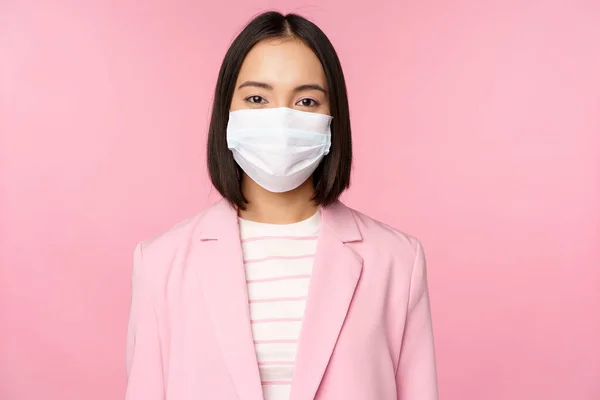 Πορτρέτο της ασιάτισσας επιχειρηματία σε ιατρική μάσκα προσώπου, φορώντας κοστούμι, έννοια της εργασίας γραφείου κατά τη διάρκεια της πανδημίας covid-19, στέκεται πάνω από ροζ φόντο — Φωτογραφία Αρχείου