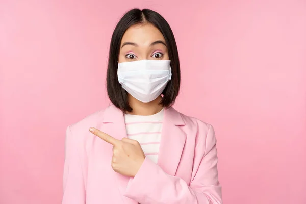 Πορτρέτο της ασιάτισσας επιχειρηματία σε ιατρική μάσκα προσώπου και κοστούμι, δείχνοντας το δάχτυλο αριστερά, δείχνει διαφήμιση, εταιρεία πανό, στούντιο ροζ φόντο — Φωτογραφία Αρχείου