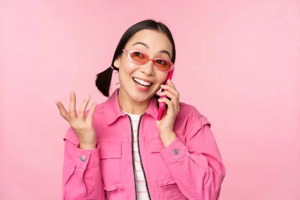 Hermosa modelo femenina coreana en gafas de sol, hablando en el teléfono móvil con la cara feliz, utilizando el servicio celular para llamar a un amigo en el teléfono inteligente, de pie sobre el fondo rosa — Foto de Stock