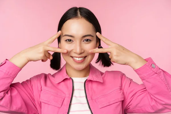 Close up of stylish asian girl smiles happy, shows peace v-sign, kawaii pose, posing against pink background — Stock Photo, Image