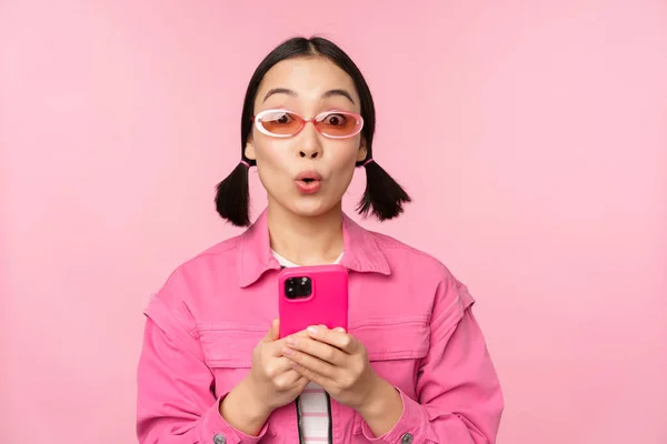 Retrato de chica coreana en gafas de sol elegantes que sostienen el teléfono móvil, utilizando la aplicación del teléfono inteligente, de pie sobre el fondo rosa — Foto de Stock