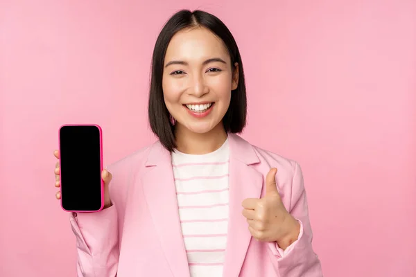 Nöjd leende asiatisk affärskvinna rekommenderar mobiltelefon app, webbplats företag på smartphone, visar skärmen och tummen upp, rosa bakgrund — Stockfoto