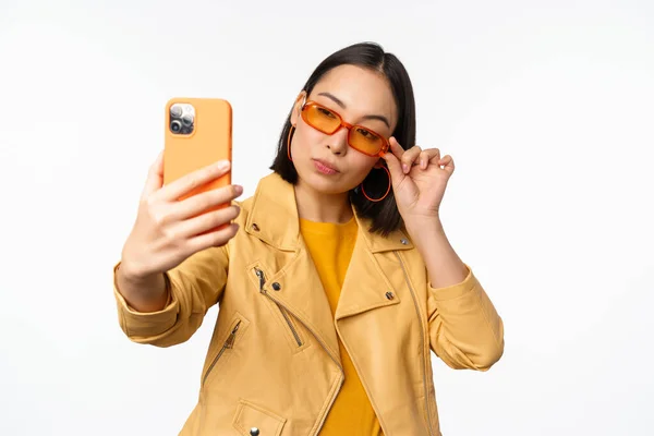 Стильная, красивая девушка в солнцезащитных очках, делающая селфи на смартфоне, позирующая для фото, держащая в руках мобильный телефон, белый студийный фон — стоковое фото