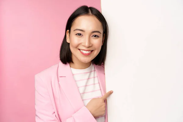 Retrato de feliz sorrindo japonês corporativo mulher apontando dedo a bordo, mostrando smth no espaço vazio, de pé em terno sobre fundo rosa — Fotografia de Stock