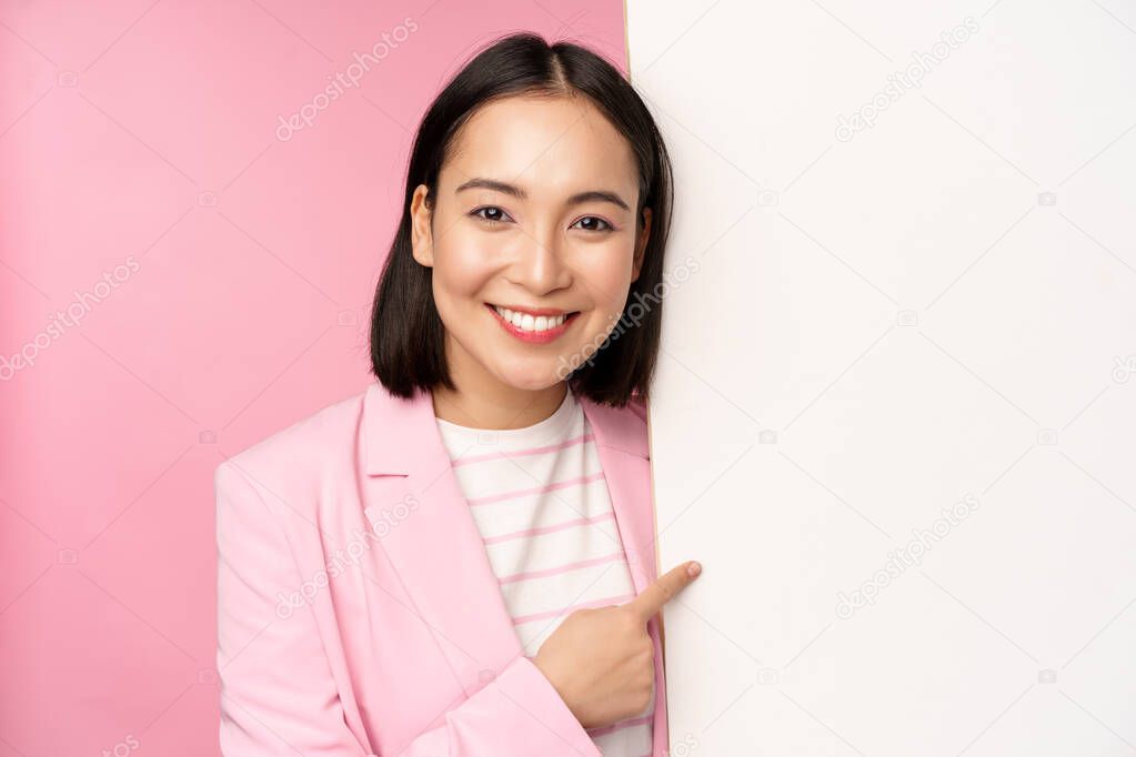 Portrait of happy smiling japanese corporate woman pointing finger at board, showing smth on empty space, standing in suit over pink background.