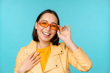 Güneş gözlüklü şık Asyalı kadın portresi, gülüyor ve gülümsüyor, mutlu görünüyor, mavi arka planda moda kıyafetlerle poz veriyor.
