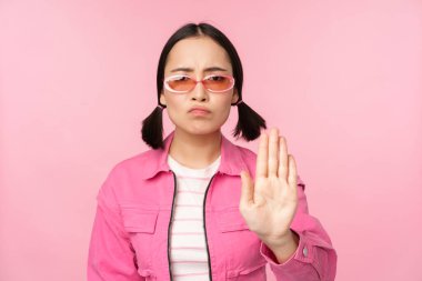 Güneş gözlüklü, ciddi, stil sahibi Asyalı bir kızın görüntüsü, durduğunu gösteriyor, jesti yasaklıyor, tabu işareti, hayır diyor, pembe arka planda duruyor.