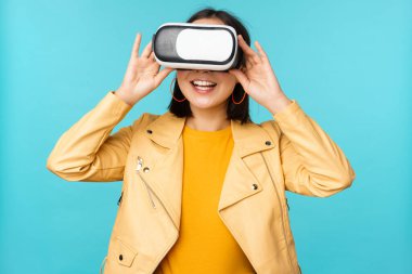 VR kulaklık takan mutlu Asyalı kadın modelin portresi. Sanal gerçeklik gözlükleriyle gülüyor ve gülümsüyor. Mavi arka planda duruyor.
