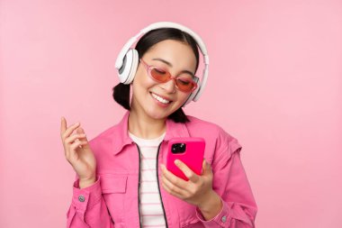 Şık Asyalı kız akıllı telefonuyla dans ediyor, kulaklıkla cep telefonu uygulamasında müzik dinliyor, gülümsüyor ve gülüyor, pembe arka planda poz veriyor.