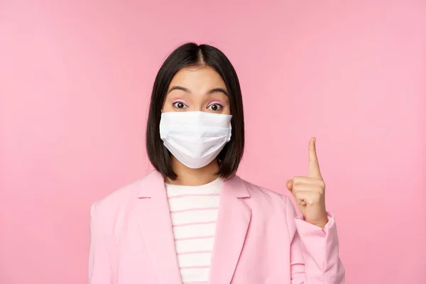 Close up retrato de mulher de negócios asiática em máscara facial médica e terno, apontando o dedo para cima, mostrando propaganda, banner superior, de pé sobre fundo rosa — Fotografia de Stock