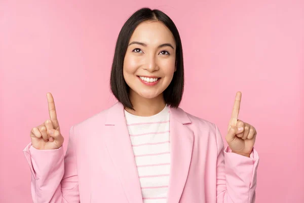 Uśmiechnięta koreańska bizneswoman, wskazująca palcem w górę, pokazująca reklamę, baner lub logo na górze, stojąca w garniturze na różowym tle — Zdjęcie stockowe