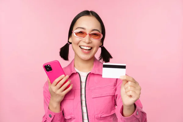 Modern vacker asiatisk flicka, skrattar och ler med mobiltelefon, kreditkort, handla online, betala med smartphone, står över rosa bakgrund — Stockfoto