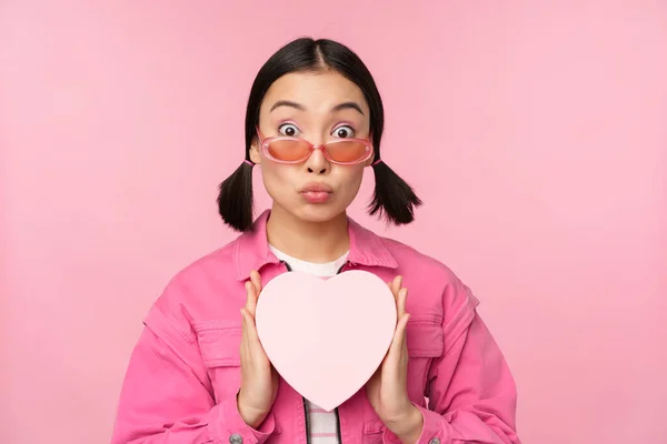 Schattig aziatisch meisje tonen hartvormige doos met cadeau, op zoek verrast en enthousiast, romantisch cadeau concept, het dragen van een zonnebril, staan over roze achtergrond — Stockfoto