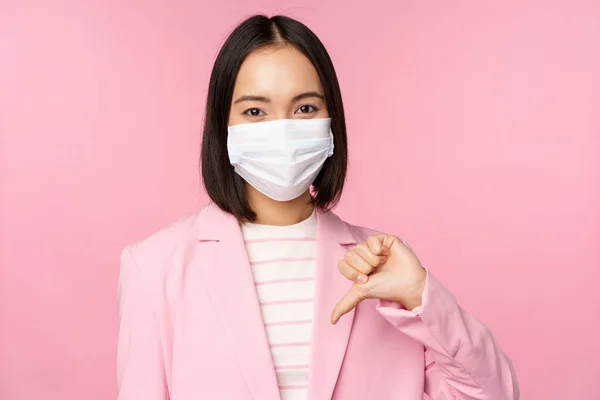 Koreańska kobieta biznesu w masce i garniturze medycznym, pokazuje kciuki w dół, nie lubi lub nie pochwala gestu, stojąc na różowym tle — Zdjęcie stockowe