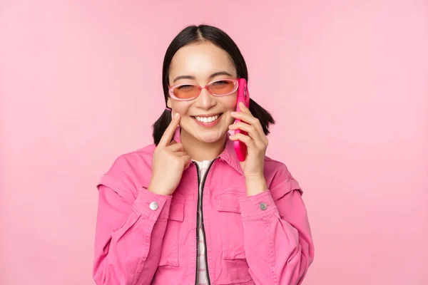 Όμορφο κορεάτικο γυναικείο μοντέλο σε γυαλιά ηλίου, μιλώντας στο κινητό τηλέφωνο με χαρούμενο πρόσωπο, χρησιμοποιώντας την υπηρεσία κινητής τηλεφωνίας για να καλέσετε τον φίλο στο smartphone, στέκεται πάνω από ροζ φόντο — Φωτογραφία Αρχείου