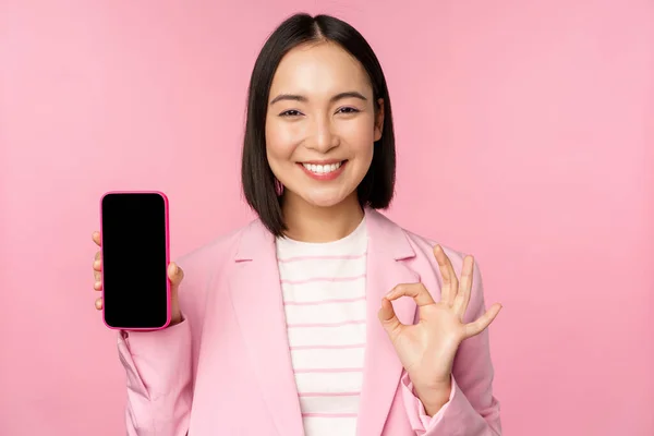 Soddisfatto sorridente donna d'affari asiatica raccomandando app per telefoni cellulari, società di siti web su smartphone, mostrando schermo e segno ok, sfondo rosa — Foto Stock