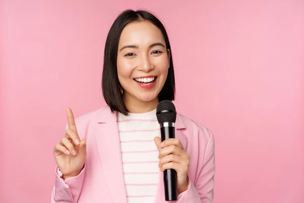 Imagen de la entusiasta mujer de negocios asiática dando discurso, hablando con micrófono, sosteniendo el micrófono, de pie en traje contra fondo de estudio rosa —  Fotos de Stock