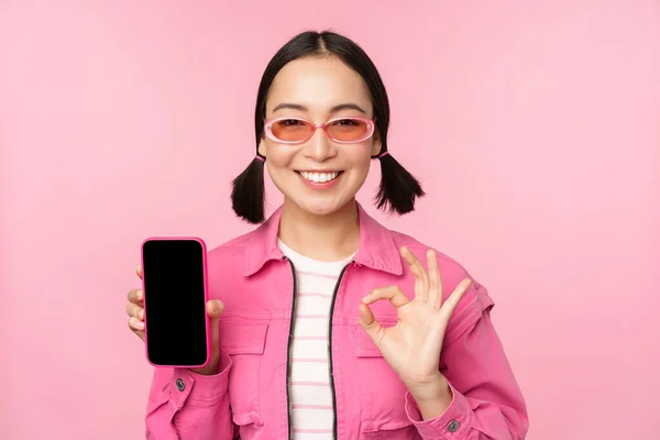 Entusiasta giovane donna asiatica mostrando ok, ok segno, sorridente soddisfatto, schermo del telefono cellulare, applicazione smartphone, in piedi su sfondo rosa — Foto Stock