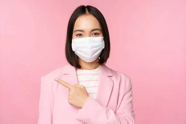 Portret azjatyckiej bizneswoman w medycznej masce i garniturze, wskazując palcem w lewo, pokazując reklamę, firmowy baner, studio różowe tło — Zdjęcie stockowe