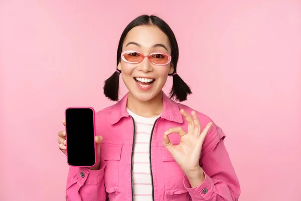 Entusiastiska unga asiatiska kvinna visar okej, ok tecken, leende nöjd, mobiltelefon skärm, smartphone ansökan, står över rosa bakgrund — Stockfoto