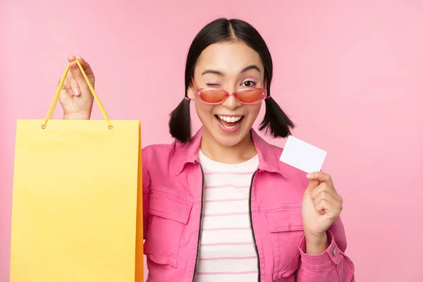 Glückliche junge asiatische Frau zeigt Kreditkarte zum Einkaufen, hält Tasche, kauft im Verkauf, geht in den Laden, speichert, steht über rosa Hintergrund — Stockfoto