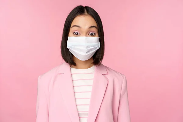 Retrato de mulher de negócios asiática em máscara facial médica, vestindo terno, conceito de trabalho de escritório durante a pandemia covid-19, de pé sobre fundo rosa — Fotografia de Stock