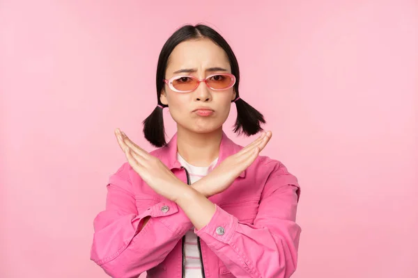 Ritratto di ragazza coreana in occhiali da sole alla moda, imbronciato deluso, mostrando stop, gesto di rifiuto, segno croce, in piedi su sfondo rosa — Foto Stock