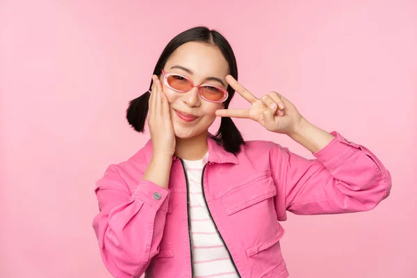 Портрет дівчини з Каваї в сонцезахисних окулярах, що показує мир, V-знак біля очей і посміхається милою в камері, виставляючи в модний одяг на рожевому фоні. — стокове фото